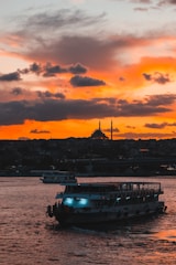 A large ferry boat travels across a body of water under a dramatic sunset sky. The vibrant orange and yellow hues contrast with the dark silhouettes of buildings and a mosque with minarets in the background.
