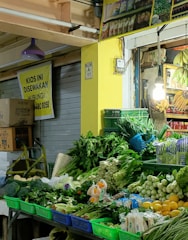 A small market stall showcasing a vibrant array of fresh vegetables and fruits. Various greens such as leafy vegetables, cucumbers, and spring onions are neatly arranged in colorful baskets. Next to the produce, bunches of bananas hang from the ceiling. The stall is illuminated by a single light bulb, adding warmth to the scene.