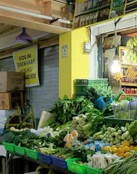 A small market stall showcasing a vibrant array of fresh vegetables and fruits. Various greens such as leafy vegetables, cucumbers, and spring onions are neatly arranged in colorful baskets. Next to the produce, bunches of bananas hang from the ceiling. The stall is illuminated by a single light bulb, adding warmth to the scene.