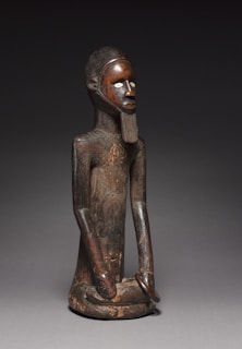 A wooden sculpture depicting a stylized human figure with elongated limbs and a prominent carved beard. The figure has an upright posture, intricately detailed facial features, and is seated with legs folded. The eyes are accentuated with a lighter material, creating a striking contrast with the dark wood. The figure's surface texture shows signs of age and wear.