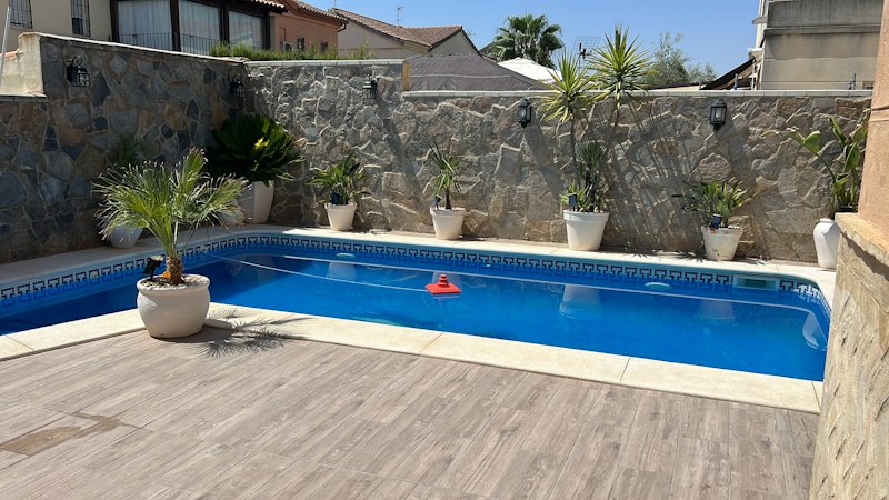A rectangular backyard pool with pale decking, white coping, and a stone wall lined with potted plants