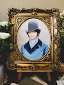 A detailed embroidery artwork features a portrait of a man wearing a vintage top hat and a blue coat. The portrait is set within an ornate, gold-colored frame. Surrounding the frame are white flowers and greenery, adding a touch of elegance.