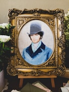 A detailed embroidery artwork features a portrait of a man wearing a vintage top hat and a blue coat. The portrait is set within an ornate, gold-colored frame. Surrounding the frame are white flowers and greenery, adding a touch of elegance.