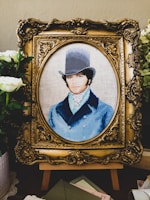 A detailed embroidery artwork features a portrait of a man wearing a vintage top hat and a blue coat. The portrait is set within an ornate, gold-colored frame. Surrounding the frame are white flowers and greenery, adding a touch of elegance.