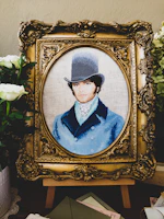 A detailed embroidery artwork features a portrait of a man wearing a vintage top hat and a blue coat. The portrait is set within an ornate, gold-colored frame. Surrounding the frame are white flowers and greenery, adding a touch of elegance.