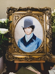 A detailed embroidery artwork features a portrait of a man wearing a vintage top hat and a blue coat. The portrait is set within an ornate, gold-colored frame. Surrounding the frame are white flowers and greenery, adding a touch of elegance.