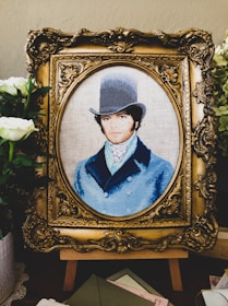 A detailed embroidery artwork features a portrait of a man wearing a vintage top hat and a blue coat. The portrait is set within an ornate, gold-colored frame. Surrounding the frame are white flowers and greenery, adding a touch of elegance.