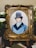 A detailed embroidery artwork features a portrait of a man wearing a vintage top hat and a blue coat. The portrait is set within an ornate, gold-colored frame. Surrounding the frame are white flowers and greenery, adding a touch of elegance.
