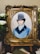 A detailed embroidery artwork features a portrait of a man wearing a vintage top hat and a blue coat. The portrait is set within an ornate, gold-colored frame. Surrounding the frame are white flowers and greenery, adding a touch of elegance.