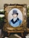 A detailed embroidery artwork features a portrait of a man wearing a vintage top hat and a blue coat. The portrait is set within an ornate, gold-colored frame. Surrounding the frame are white flowers and greenery, adding a touch of elegance.
