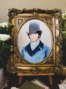 A detailed embroidery artwork features a portrait of a man wearing a vintage top hat and a blue coat. The portrait is set within an ornate, gold-colored frame. Surrounding the frame are white flowers and greenery, adding a touch of elegance.