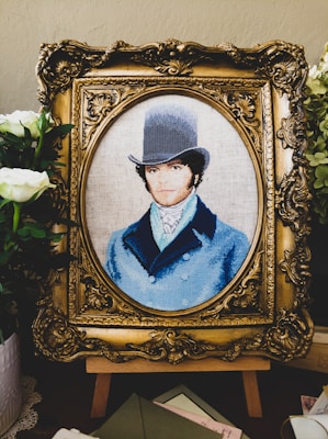 A detailed embroidery artwork features a portrait of a man wearing a vintage top hat and a blue coat. The portrait is set within an ornate, gold-colored frame. Surrounding the frame are white flowers and greenery, adding a touch of elegance.