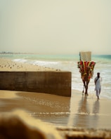 A man in traditional attire walks alongside a decorated camel on a sandy beach. The camel is adorned with colorful decorations, and the beach is lined with scattered rocks and a concrete barrier. The ocean waves gently crash onto the shore under a hazy sky.