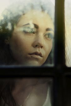 A woman gazes pensively through a glass pane, with soft light casting a gentle glow on her face. Her expression suggests contemplation or introspection, and the window is slightly blurred with natural light creating subtle reflections.