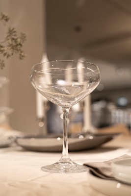 A clear, elegant cocktail glass sits on a neatly set table with plates and napkins. Soft focus on the background highlights a minimalistic dining setup, lit by warm lighting, creating a sophisticated atmosphere.