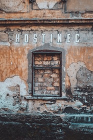 A weathered, old building facade with a blocked-up window made of bricks and surrounded by peeling, faded paint in earthy tones. The word 'HOSTINEC' is prominently displayed above the window in aged letters.