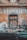 A weathered, old building facade with a blocked-up window made of bricks and surrounded by peeling, faded paint in earthy tones. The word 'HOSTINEC' is prominently displayed above the window in aged letters.