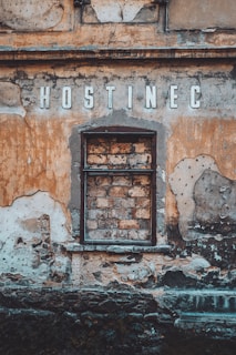A weathered, old building facade with a blocked-up window made of bricks and surrounded by peeling, faded paint in earthy tones. The word 'HOSTINEC' is prominently displayed above the window in aged letters.