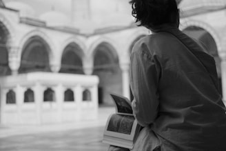 A person with short hair is sitting and holding a large open book, gazing at an architectural structure. The setting appears to be an outdoor courtyard featuring arches and textured stonework. The image is in black and white, adding a contemplative and timeless feel.