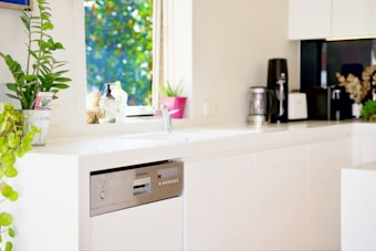 A modern kitchen counter with a minimalistic design features white cabinetry and a built-in dishwasher. On the counter are a coffee maker, a toaster, and a variety of potted plants, including a tall green plant and a smaller plant in a pink pot. A window with a view of green foliage lets in natural light.