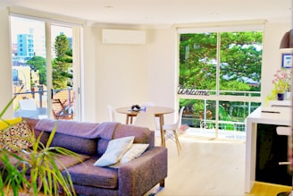 A bright and cozy living room with a large gray sofa adorned with patterned pillows. A round wooden dining table with two modern chairs is near a large window, offering a view of greenery outside. The room is decorated with houseplants and there is a 'Welcome' sign on the window. A contemporary kitchen counter is visible with some potted flowers placed on it.