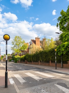 A tree-lined suburban street featuring a crosswalk and classic architecture. The brick buildings have tall chimneys and are partially obscured by lush green hedges. Overhead, a blue sky is dotted with fluffy white clouds, contributing to a serene and pleasant atmosphere.