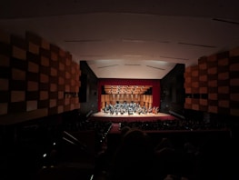 An auditorium featuring a symphony orchestra performing on stage with musicians in formal attire. The stage is illuminated, showcasing a wooden backdrop and a variety of instruments. The large hall is filled with an audience in a dimly lit area.