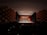 An auditorium featuring a symphony orchestra performing on stage with musicians in formal attire. The stage is illuminated, showcasing a wooden backdrop and a variety of instruments. The large hall is filled with an audience in a dimly lit area.