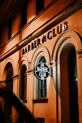 A warmly lit facade of a building featuring an illuminated sign with the text 'Barber Club' in a stylish font. The architecture includes arched windows and a classic design, contributing to a vintage and inviting atmosphere.