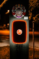 A modern electric vehicle charging station is illuminated at night. The charger has bold red lighting outlining its frame. Above the charger, there is a detailed and colorful coat of arms. The background is dimly lit, suggesting it's a suburban area or a park.