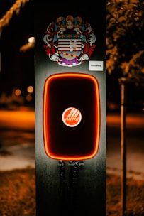 A modern electric vehicle charging station is illuminated at night. The charger has bold red lighting outlining its frame. Above the charger, there is a detailed and colorful coat of arms. The background is dimly lit, suggesting it's a suburban area or a park.