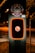 A modern electric vehicle charging station is illuminated at night. The charger has bold red lighting outlining its frame. Above the charger, there is a detailed and colorful coat of arms. The background is dimly lit, suggesting it's a suburban area or a park.