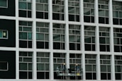 Two construction workers in hard hats stand on a suspended platform, cleaning the windows of a tall building that features a grid-like pattern of glass panes and white frames.