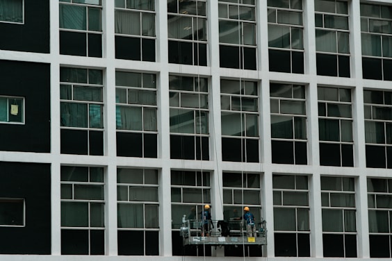 Two construction workers in hard hats stand on a suspended platform, cleaning the windows of a tall building that features a grid-like pattern of glass panes and white frames.