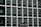 Two construction workers in hard hats stand on a suspended platform, cleaning the windows of a tall building that features a grid-like pattern of glass panes and white frames.