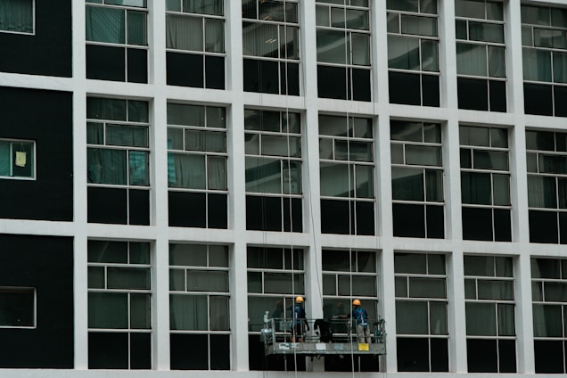 Two construction workers in hard hats stand on a suspended platform, cleaning the windows of a tall building that features a grid-like pattern of glass panes and white frames.