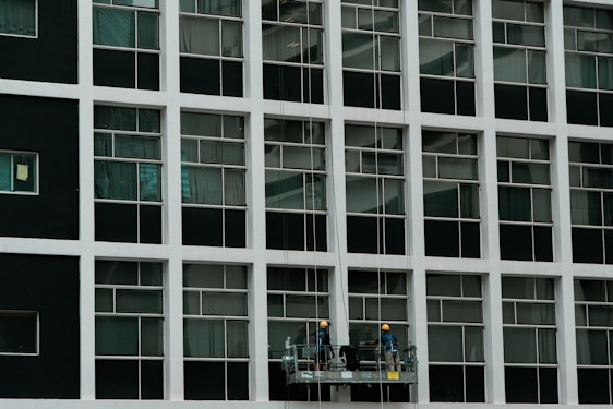 Two construction workers in hard hats stand on a suspended platform, cleaning the windows of a tall building that features a grid-like pattern of glass panes and white frames.