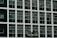 Two construction workers in hard hats stand on a suspended platform, cleaning the windows of a tall building that features a grid-like pattern of glass panes and white frames.
