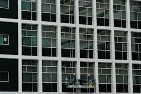 Two construction workers in hard hats stand on a suspended platform, cleaning the windows of a tall building that features a grid-like pattern of glass panes and white frames.