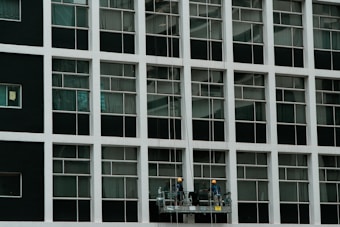 Two construction workers in hard hats stand on a suspended platform, cleaning the windows of a tall building that features a grid-like pattern of glass panes and white frames.