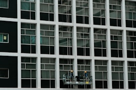 Two construction workers in hard hats stand on a suspended platform, cleaning the windows of a tall building that features a grid-like pattern of glass panes and white frames.