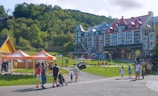 A scenic area with colorful, multi-story buildings in a European architectural style, set against a lush green hillside. The foreground features people walking and standing near orange tents labeled 'Ziptrek'. A small playground and water play area are visible in front of the buildings, with families and children enjoying the space.