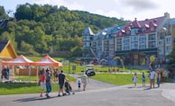 A scenic area with colorful, multi-story buildings in a European architectural style, set against a lush green hillside. The foreground features people walking and standing near orange tents labeled 'Ziptrek'. A small playground and water play area are visible in front of the buildings, with families and children enjoying the space.