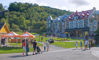 A scenic area with colorful, multi-story buildings in a European architectural style, set against a lush green hillside. The foreground features people walking and standing near orange tents labeled 'Ziptrek'. A small playground and water play area are visible in front of the buildings, with families and children enjoying the space.