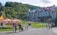 A scenic area with colorful, multi-story buildings in a European architectural style, set against a lush green hillside. The foreground features people walking and standing near orange tents labeled 'Ziptrek'. A small playground and water play area are visible in front of the buildings, with families and children enjoying the space.