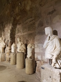 Several ancient stone statues are displayed along a rustic stone wall, each set upon a cylindrical pedestal. The statues are mostly headless, showcasing intricate drapery and detailed sculpting. The setting appears to be a historic gallery or museum with dim lighting, highlighting the textures of the stone.