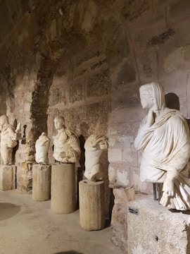 Several ancient stone statues are displayed along a rustic stone wall, each set upon a cylindrical pedestal. The statues are mostly headless, showcasing intricate drapery and detailed sculpting. The setting appears to be a historic gallery or museum with dim lighting, highlighting the textures of the stone.