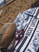 On a sandy beach, a patterned towel with the text 'WEST COAST' is spread out. A pair of red sunglasses and a woven bag with straps are placed on the towel. The ocean is visible in the background with some people and beach chairs near the shore.