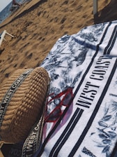 On a sandy beach, a patterned towel with the text 'WEST COAST' is spread out. A pair of red sunglasses and a woven bag with straps are placed on the towel. The ocean is visible in the background with some people and beach chairs near the shore.