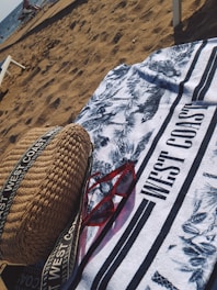 On a sandy beach, a patterned towel with the text 'WEST COAST' is spread out. A pair of red sunglasses and a woven bag with straps are placed on the towel. The ocean is visible in the background with some people and beach chairs near the shore.